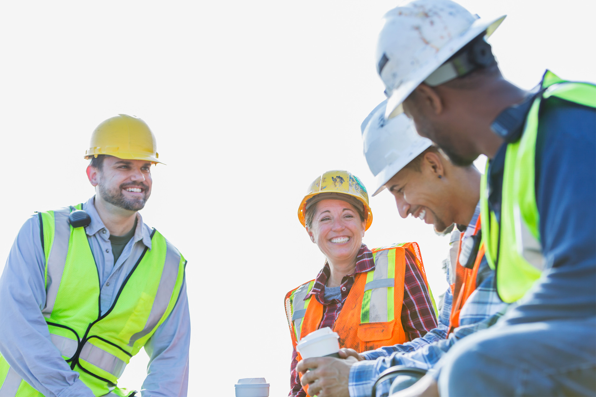 A multi-ethnic group of workers, a woman and three men, wearing safety vests and hardhats taking a coffee break.